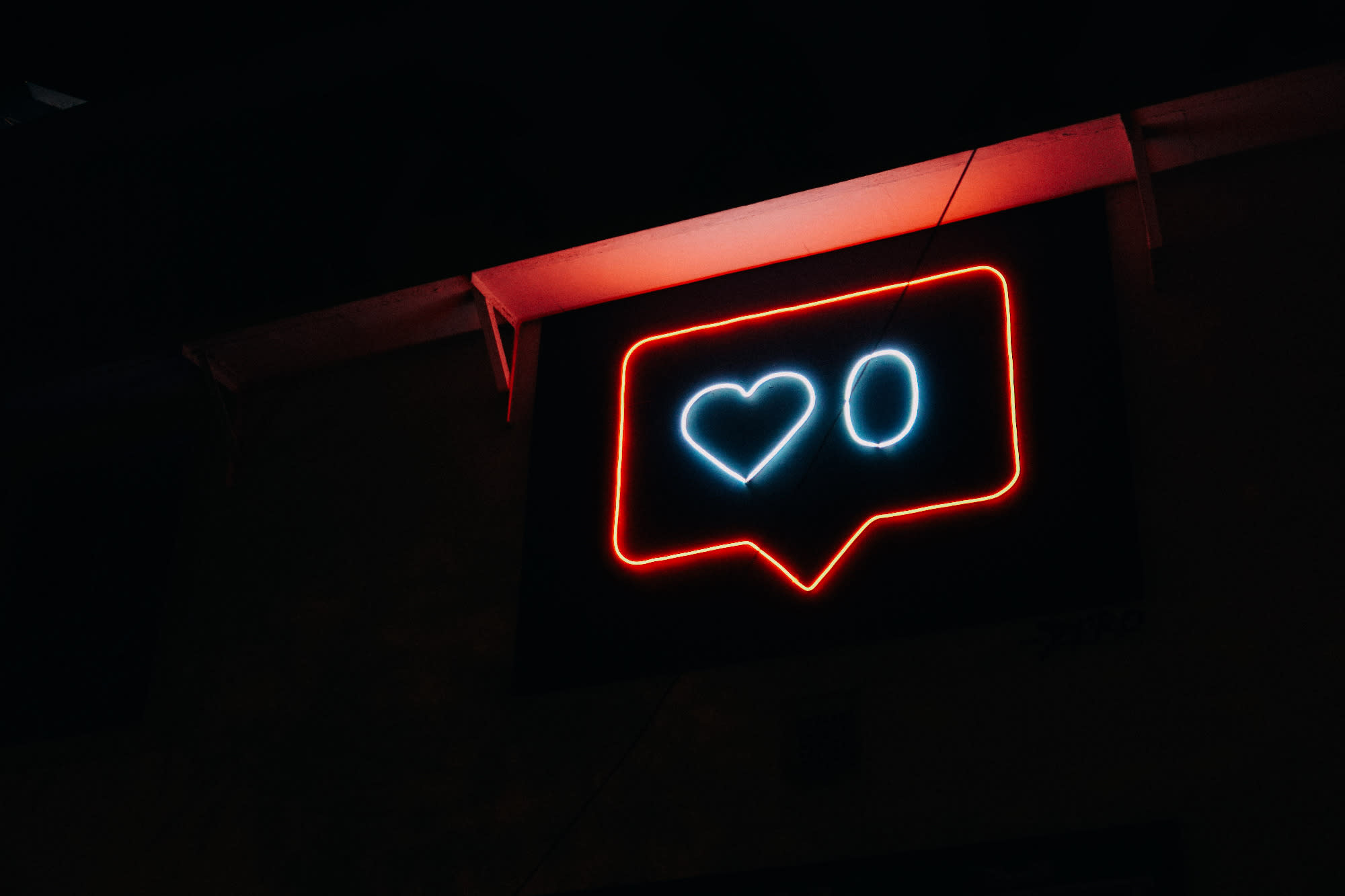 A red neon light showing a speech bubble with a heart in it and a 0 - denoting no likes on social media in a black background - that looks like nighttime
