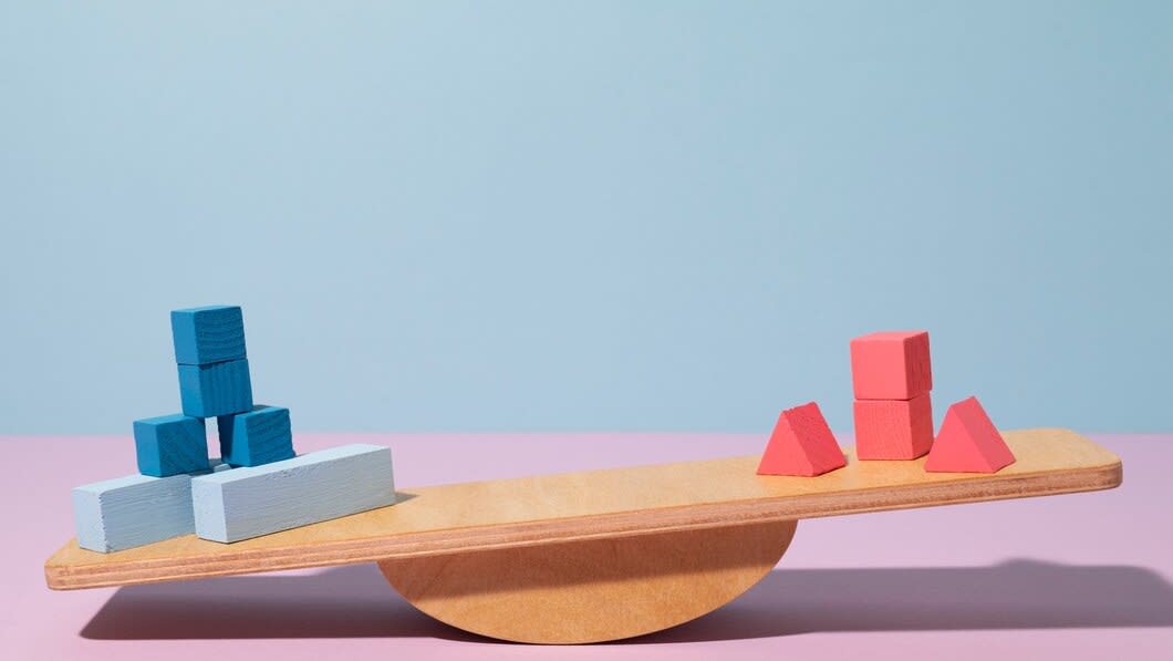 A wooden seesaw with coloured blocks on it. Blue building blocks on one end (bottom of seesaw) and red blocks on the other end.