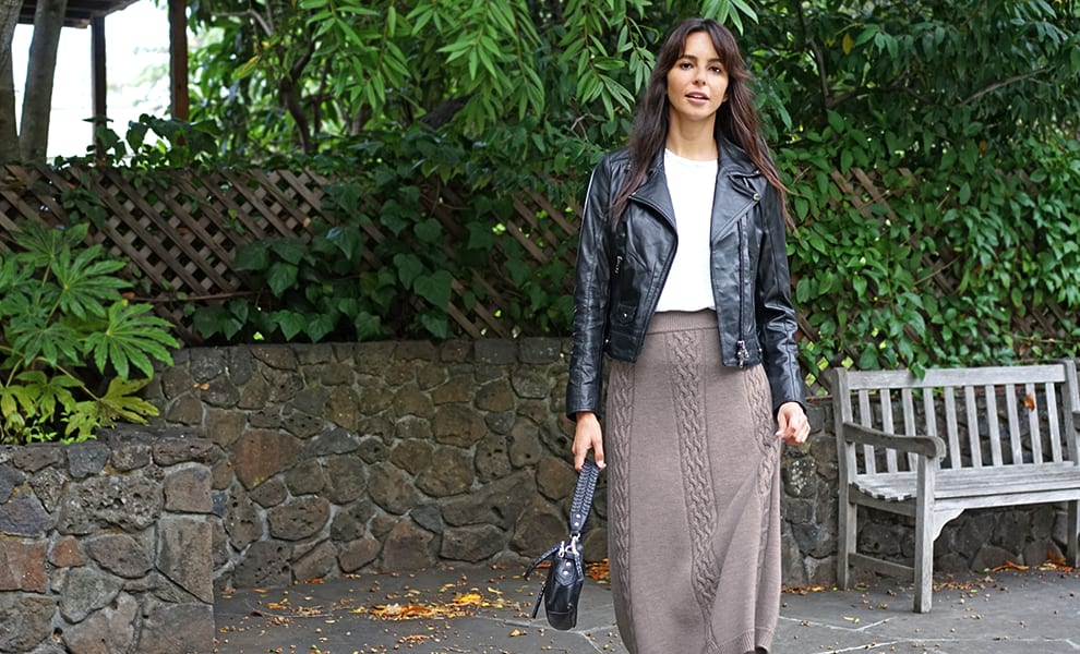 Woman wearing a leather jacket and grey knit skirt, standing in front of a leafy green background.