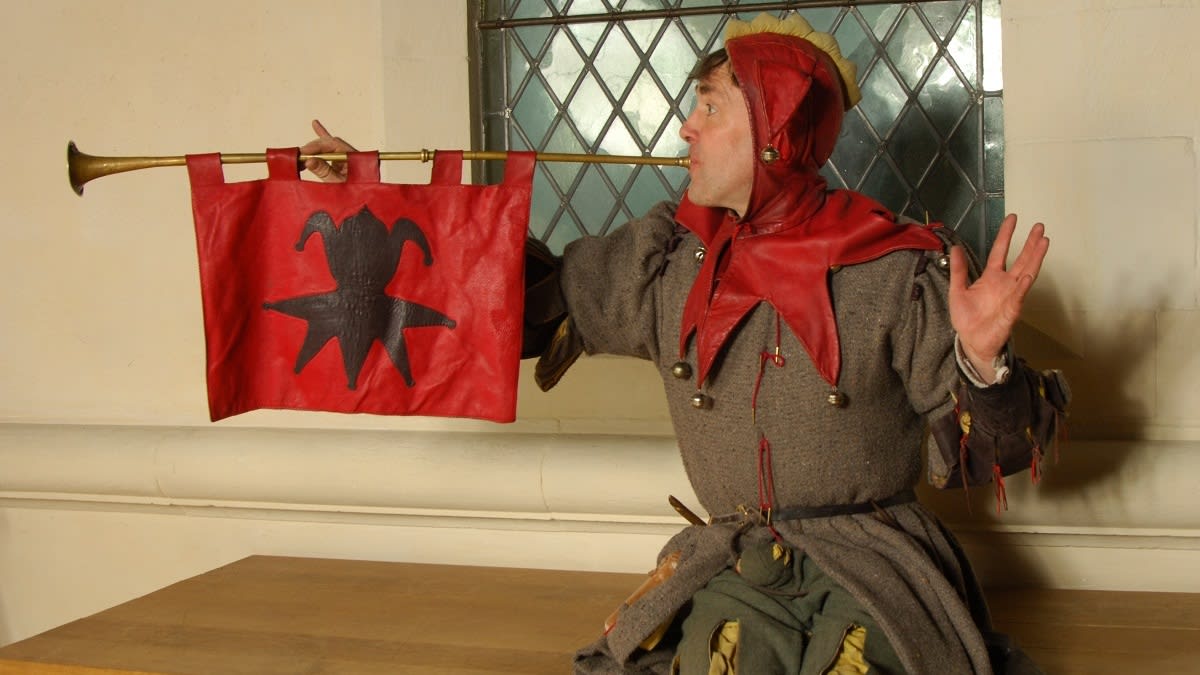 A reenactor dressed as a court jester blows into a large horn or trumpet