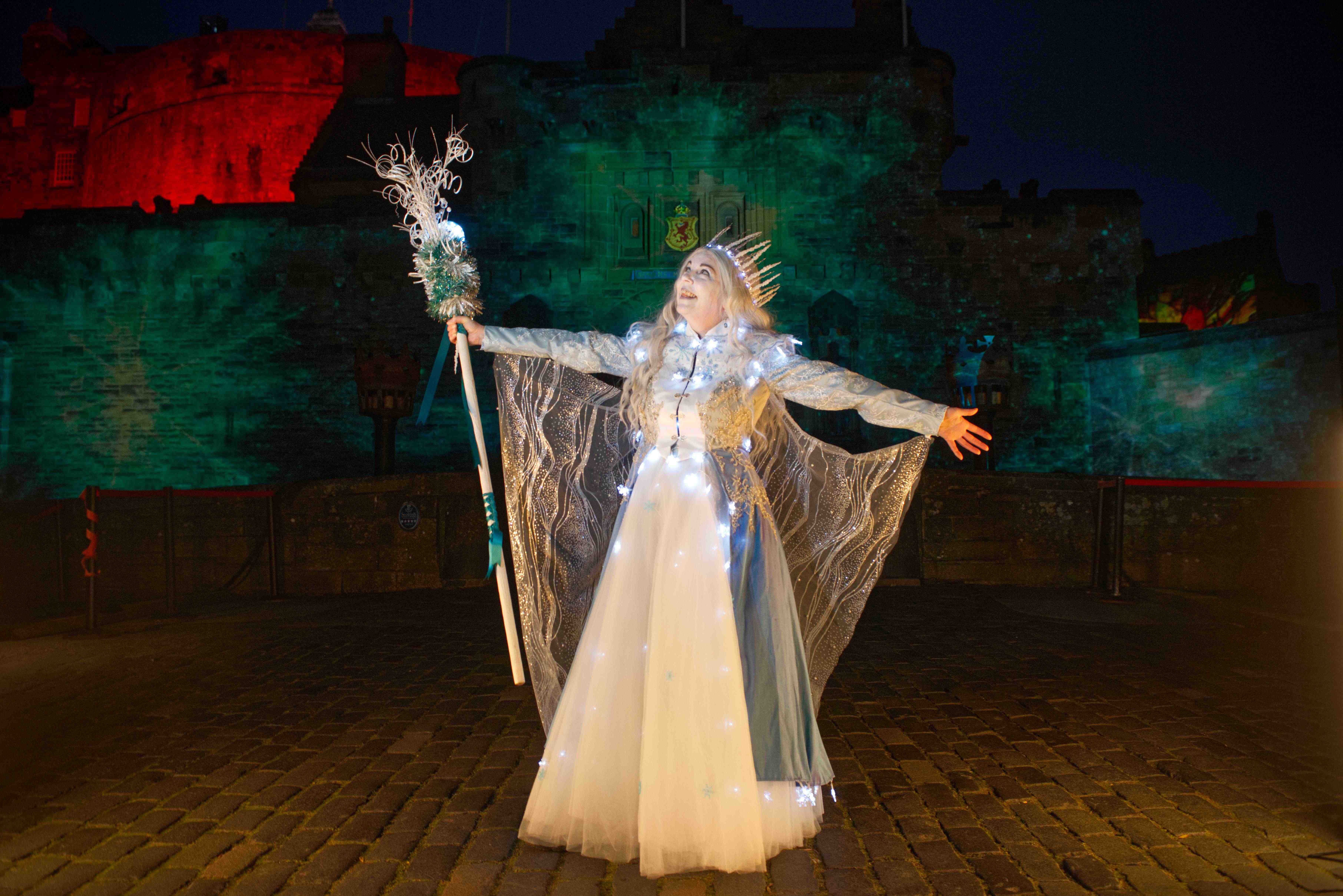 Woman in bright Snow Queen costume gestures in front of a castle entrance floodlit in bright lights