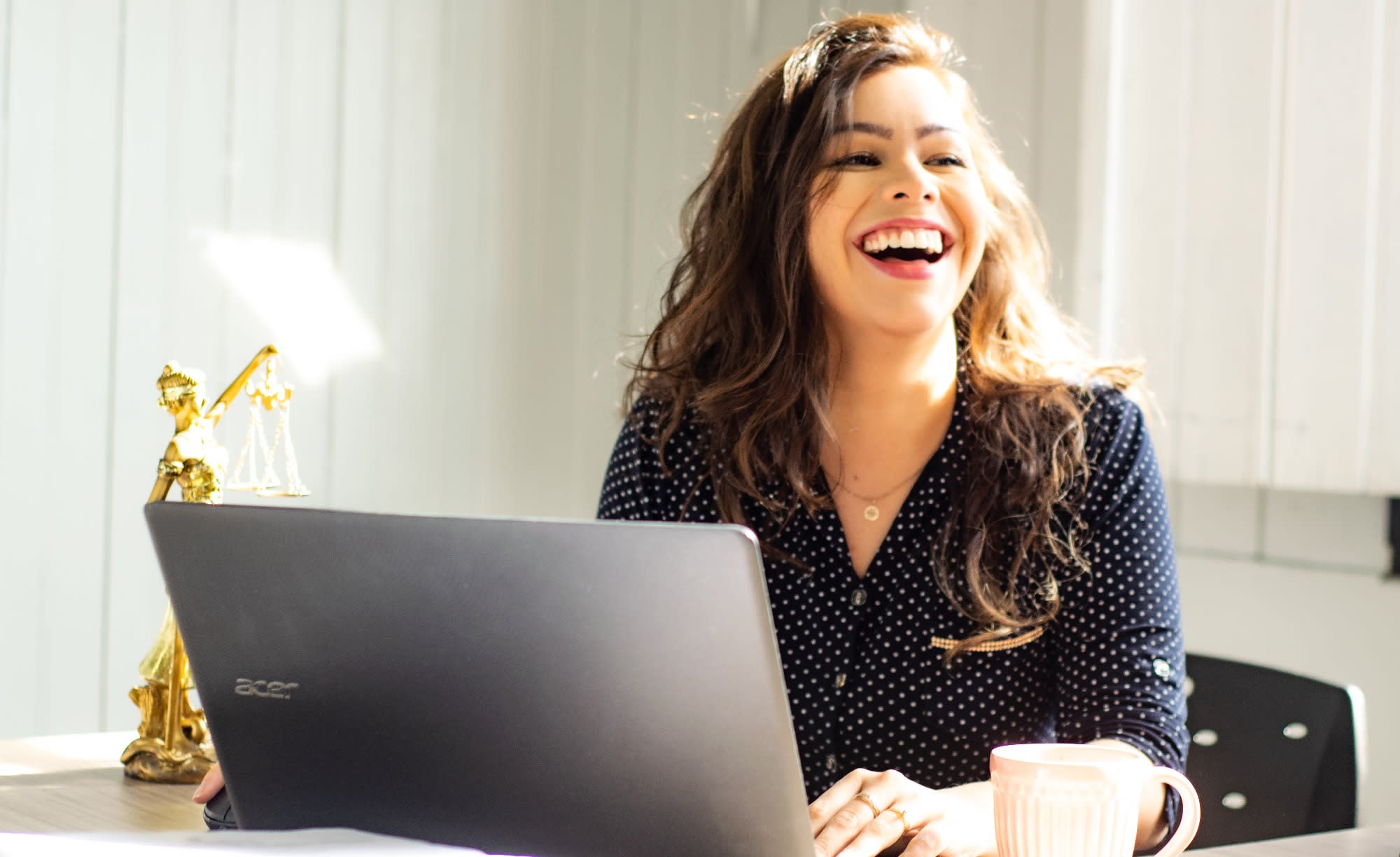 White woman with long dark hair and and a blue dress with white polka dots, is laughing in front of a laptop. To her side is a scales of justice.