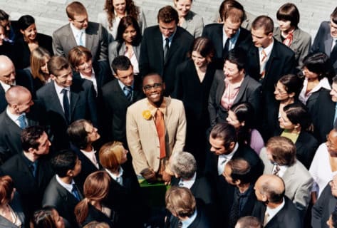 A man standing, smiling, and looking up at the camera while a crowd of people surround him and stare
