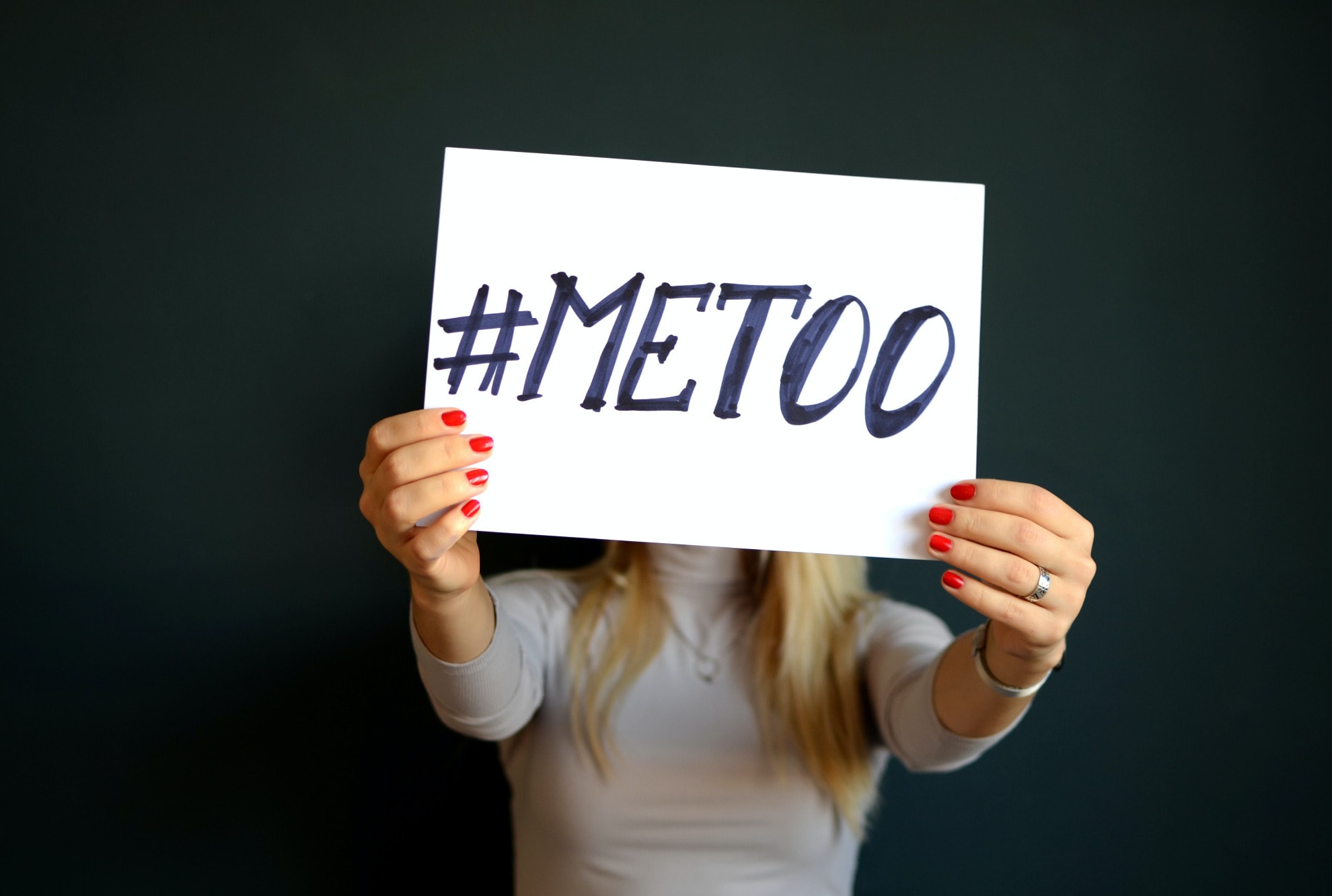 White blonde woman holding up a sign with #MeToo on it in front of her face. There is a black background.