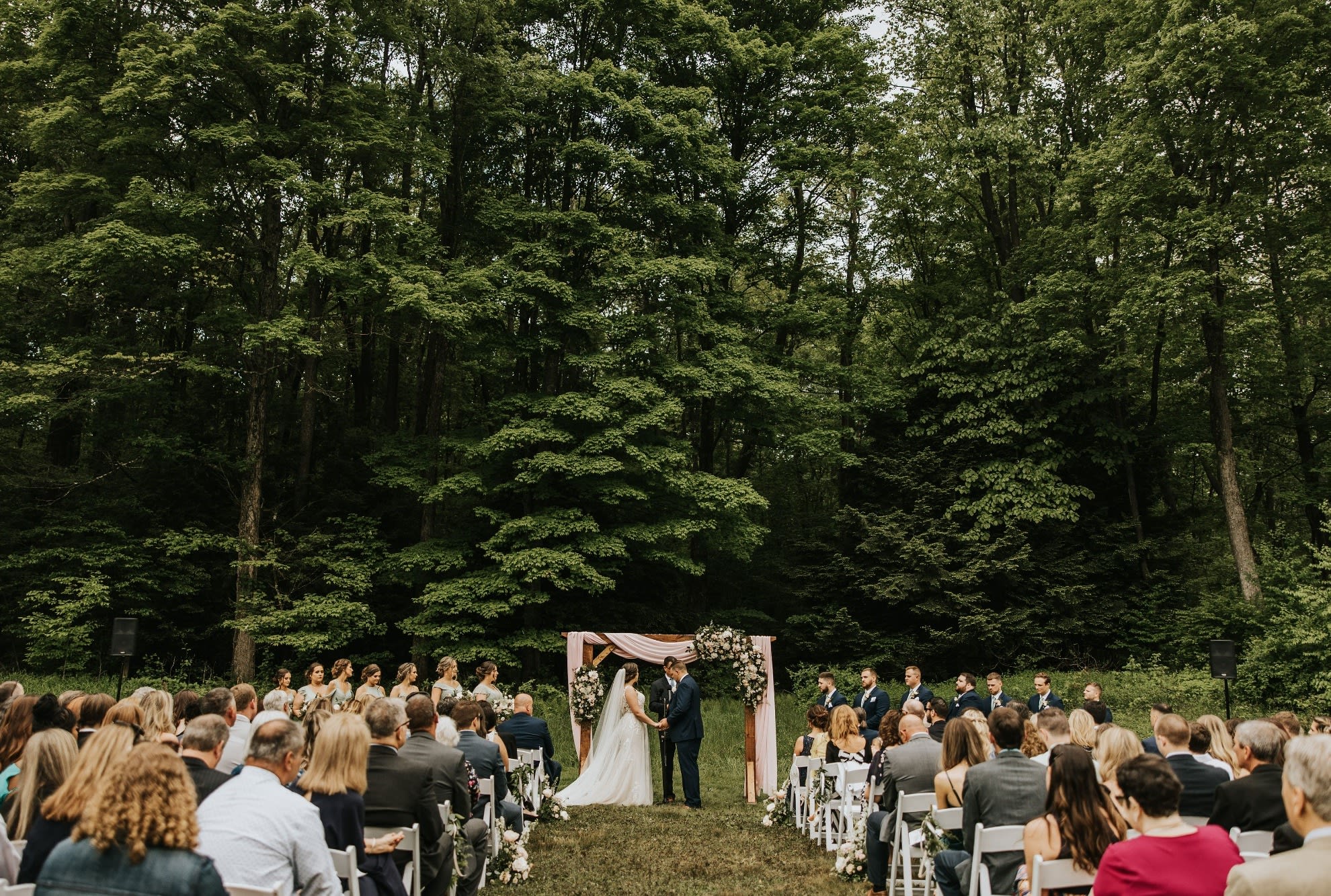 Bride and  groom stand under an arch holding hands as they recite their vows while a large group of friends and family watch from white garden chairs.