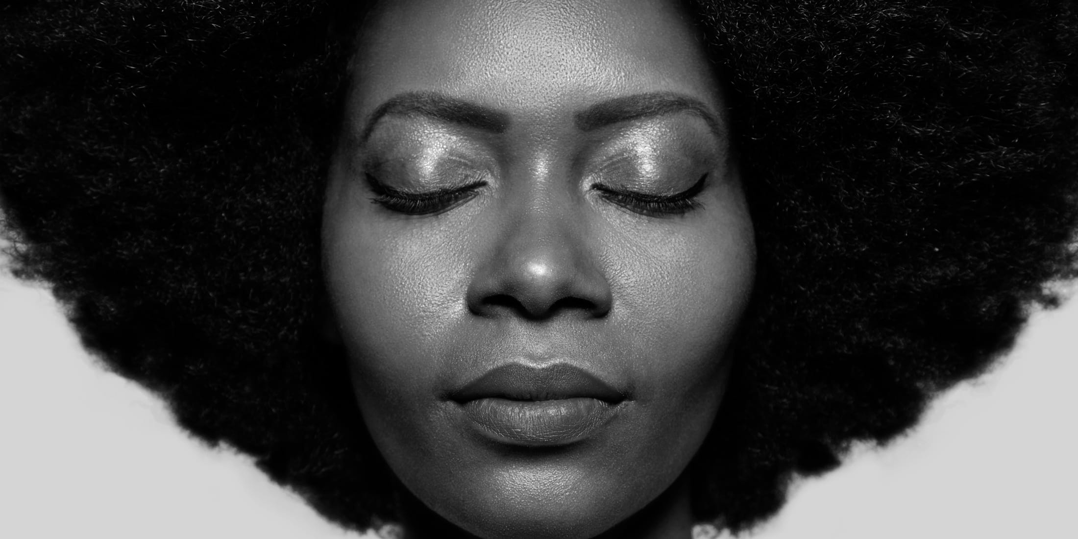 A photograph of a Black woman with an afro and her eyes closed. Her skin glistens. She smiles softly. And also, her eyebrows are beautifully shaped.
