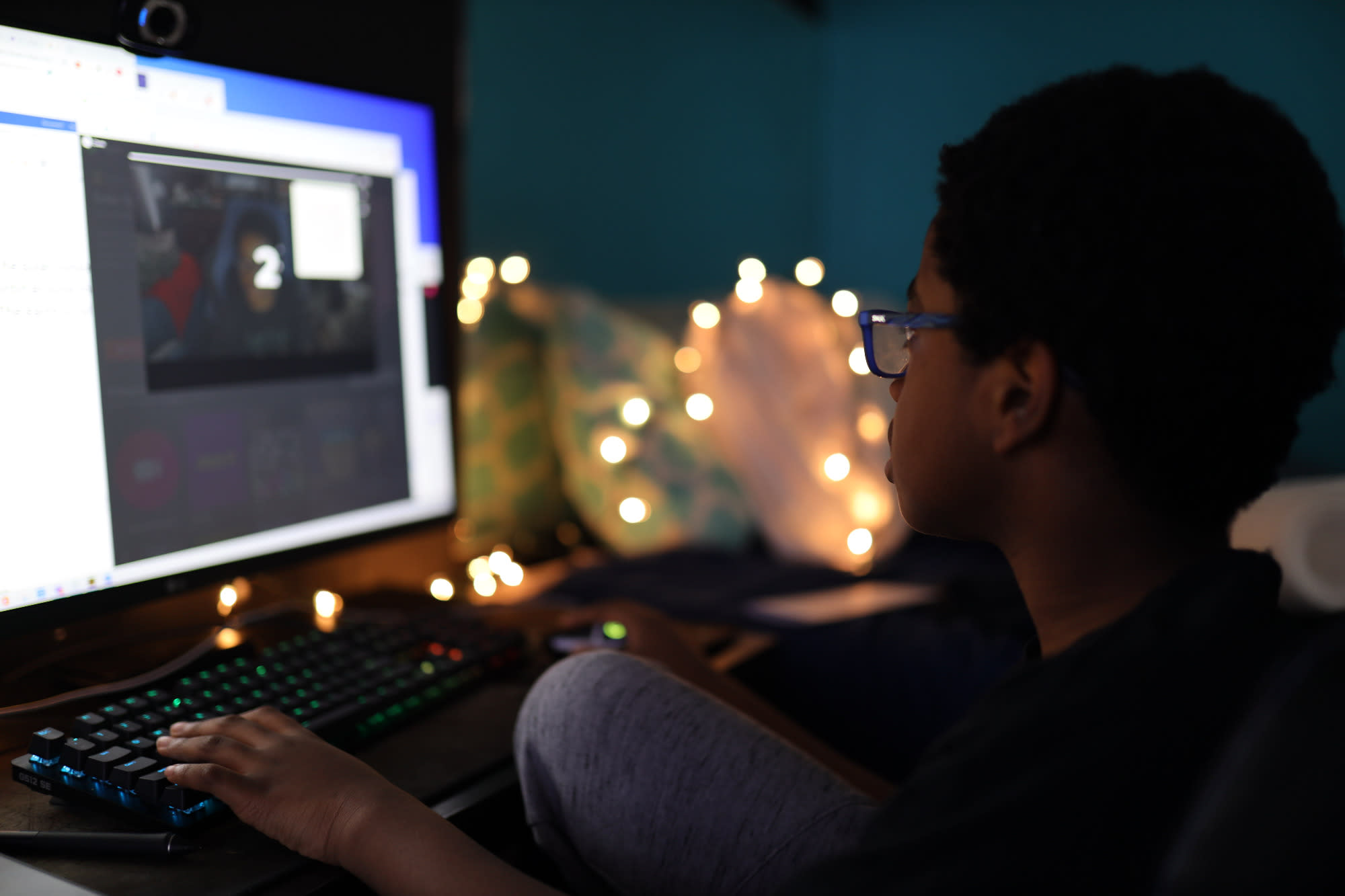 A Black teenager is playing computer games in the dark. In the background are fairylights