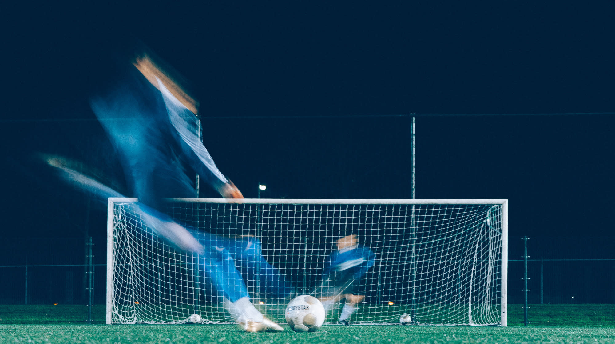 A photo on a long release so that the figures are blurred as they move. The shot is of a footballer shooting a penalty into the goal. It is at night. 