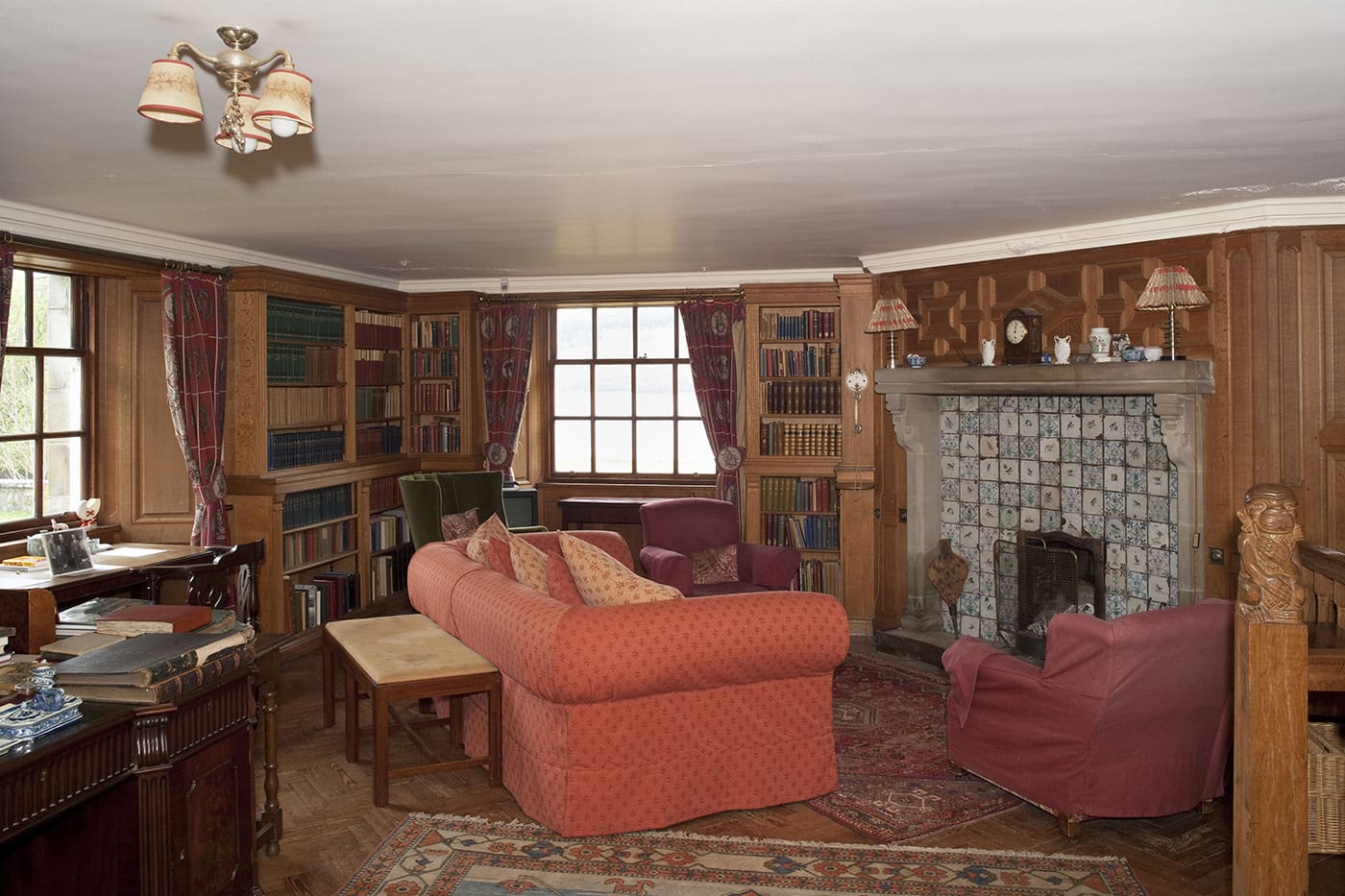 A living room with wood paneled walls, sofas, wooden desks, a fireplace and filled bookcases lining the walls