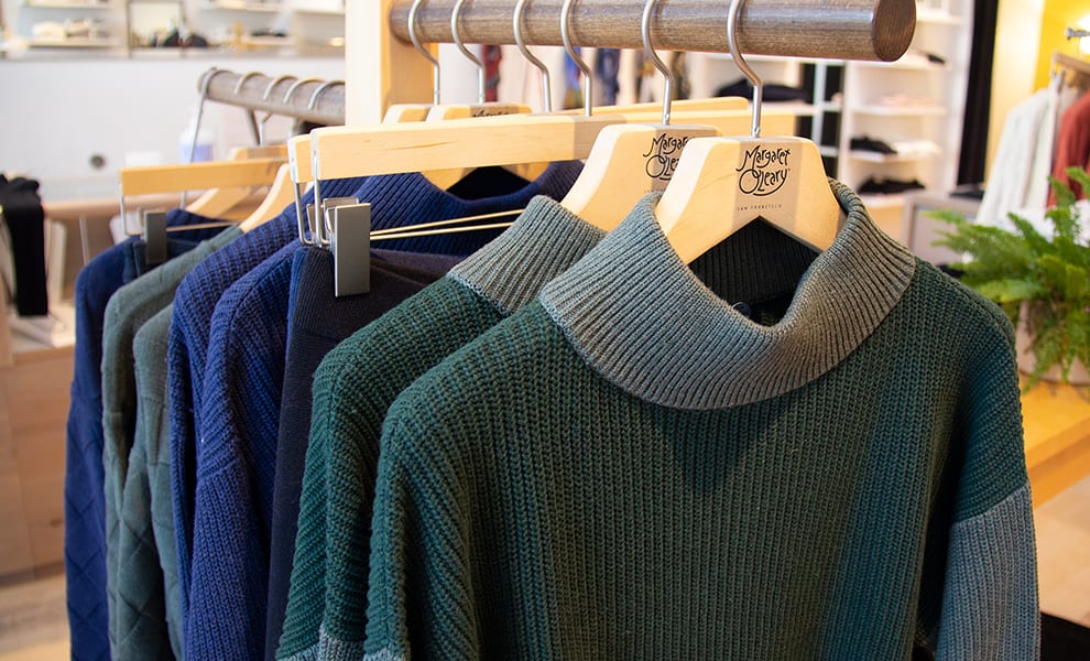 Close up view of green knit sweaters hung on wooden hangers on a small rack inside a boutique store. 