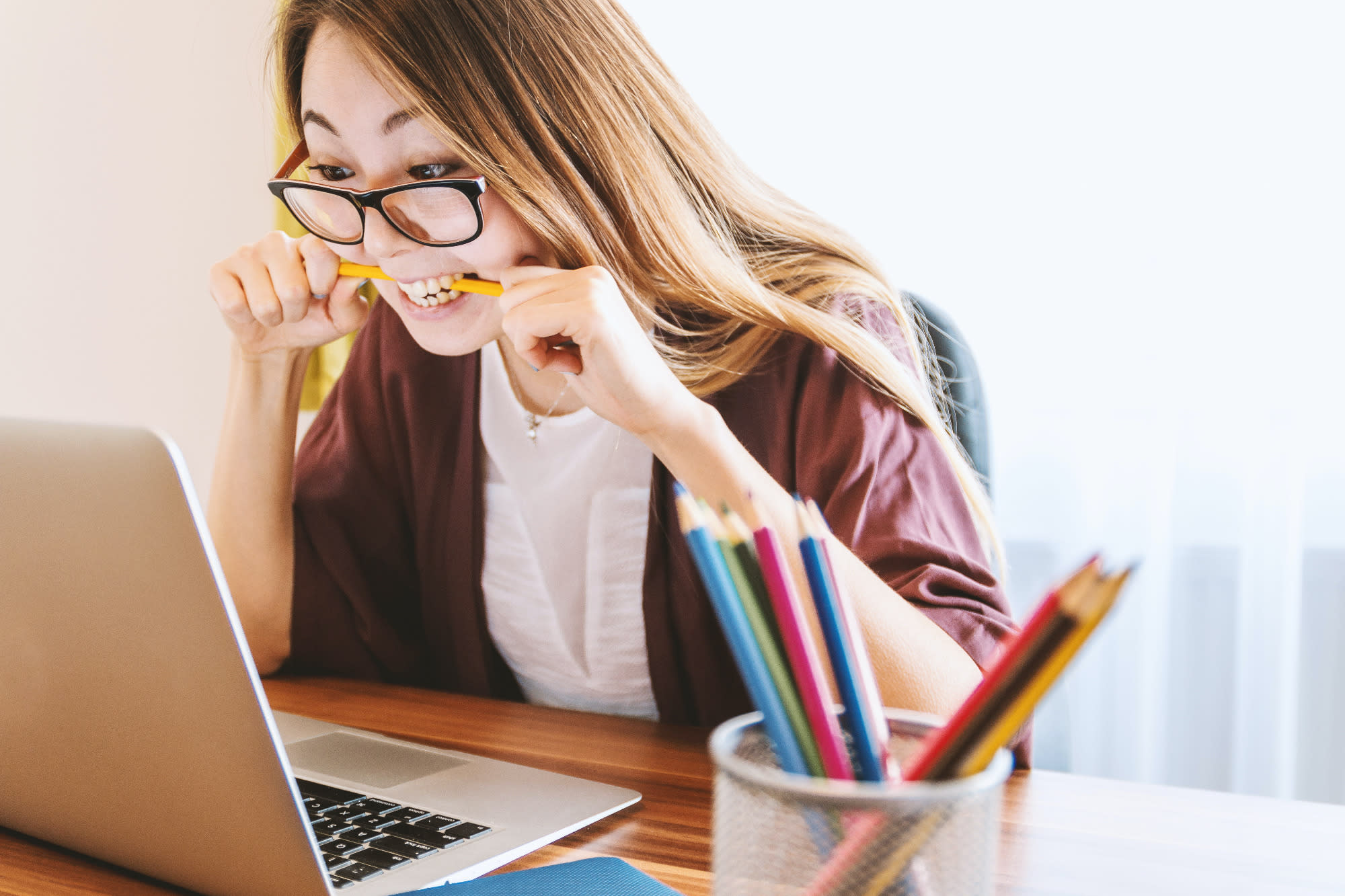 An Asian young woman in glasses and long loose dark hair, studying her laptop intently and biting on a pencil. To the right of the picture is a jar with more pencils 