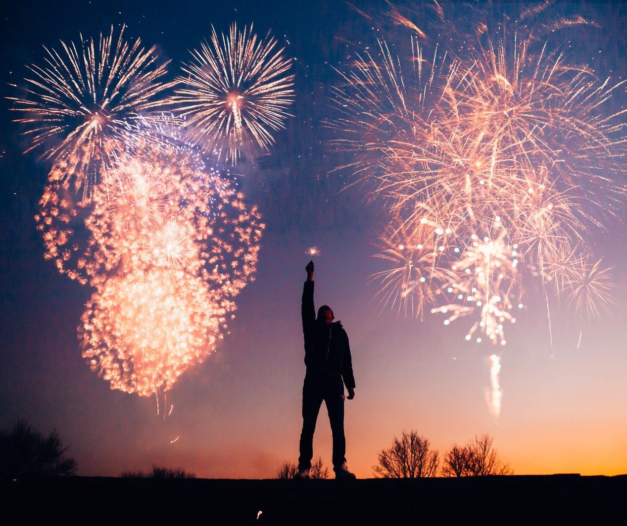 A person silhouetted against a peach and purple sunset raises their hand with a lit sparkler
