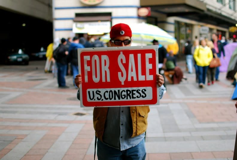 A protestor standing holding a sign up saying 'For Sale US Congress' from the Seattle Occupy protests, in the background are groups of protestors