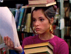 Librarian walking through library with books on head and reading from papers.