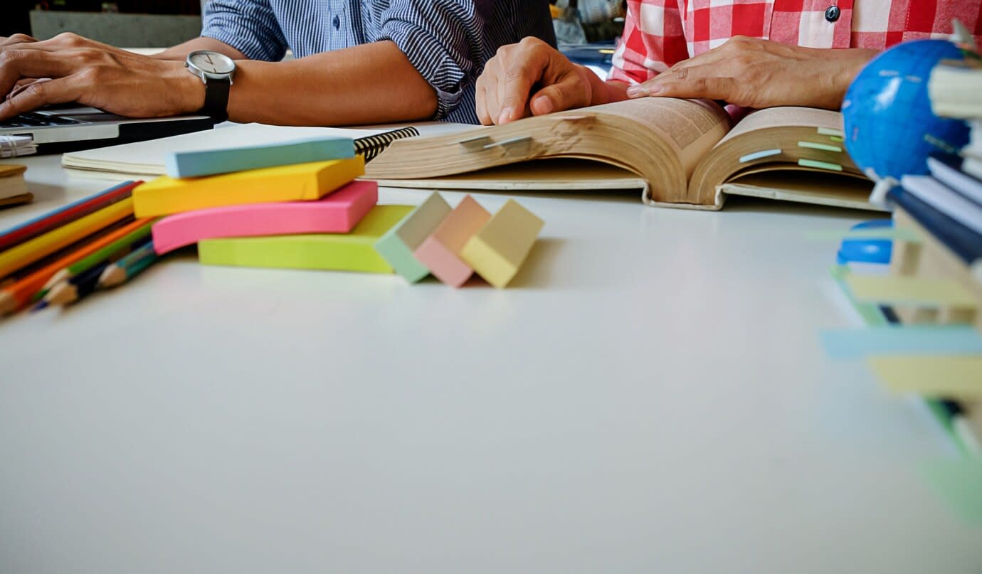 Two people sitting side by side at a desk. Only their hands and part of their upper body are visible. One os typing and the other resting their hands on an open book. Post-it notes, pencils and a small globe are at the foreground of the image. and 