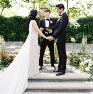 A bride and groom stand holding hands in front of an officiant reciting their vows on their wedding day