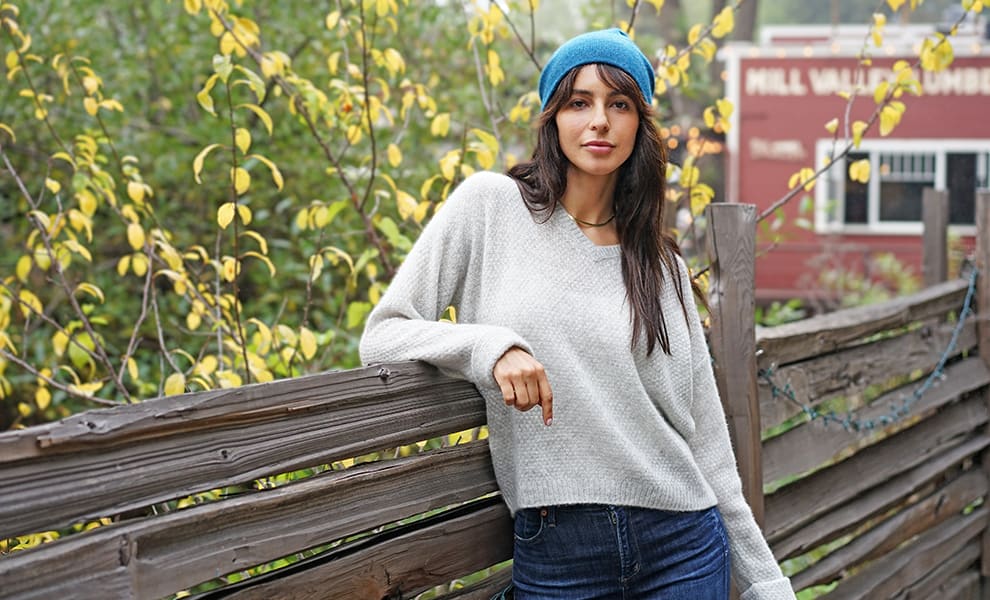 A woman in a grey sweater and blue beanie leans against a wooden fence.
