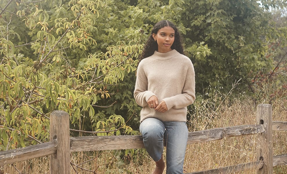 A woman in a beige turtle neck sweaters sits atop a wooden fence in a grassy field.