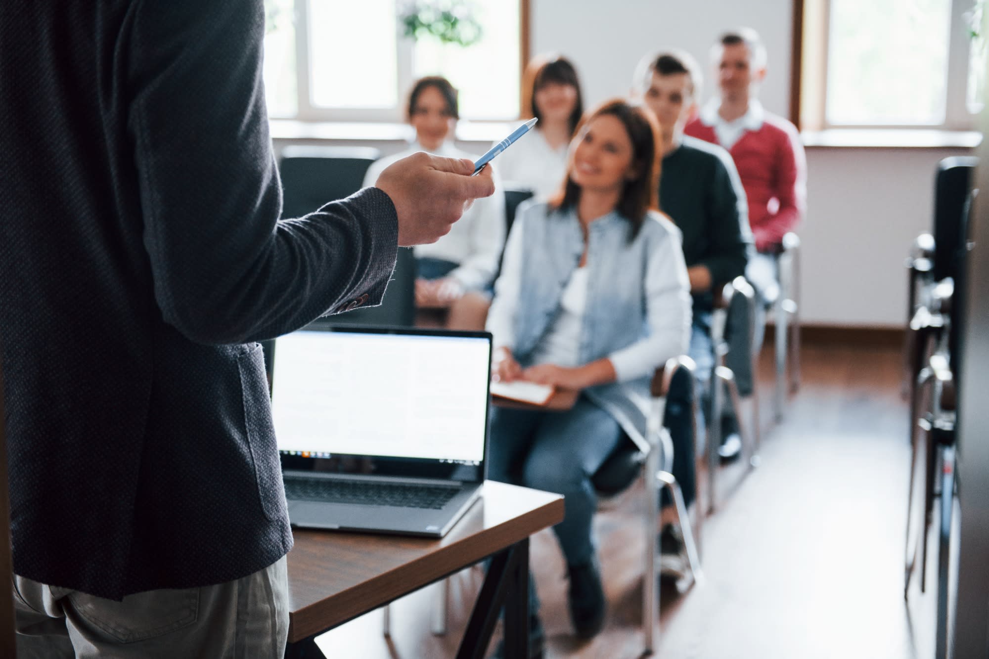 A lecture taking place in a classroom with adults. The back and right arm of the lecturer, standing behind a desk with an open laptop, are visible. He is holding a pen and facing the audience. Five smiling people are sitting down and facing him. 