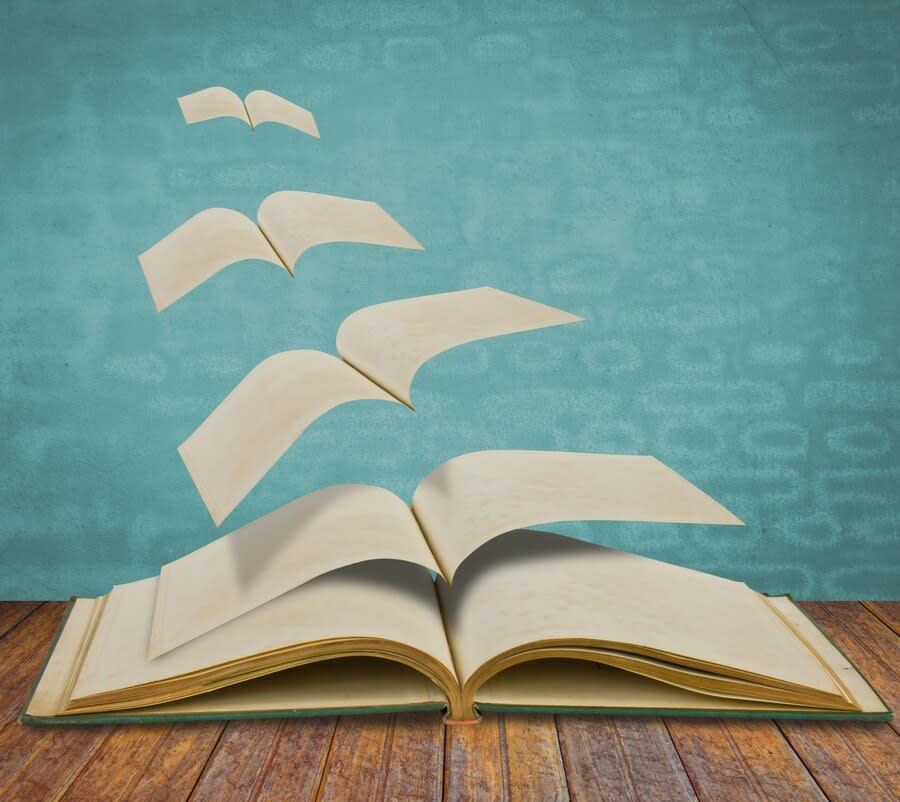 An open book on a wooden surface against a blue background. Several pages from the open book fly upwards.