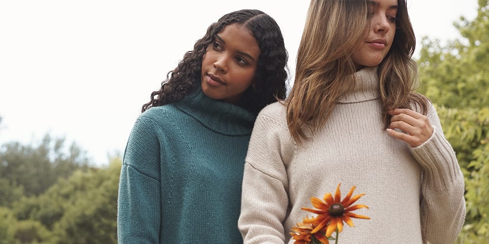 Two women in turtle neck sweaters stand in a grassy field holding an orange flower.