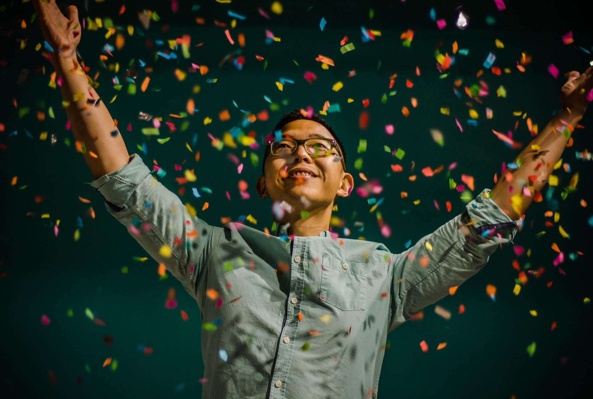 An Asian person in a grey shirt raises their hands and smiles, surrounded by a shower of confetti