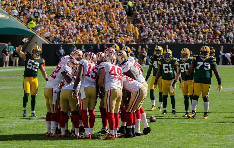 The San Francisco 49ers in a huddle on a football field. Crowds in the background. 