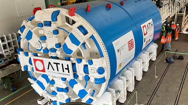 Eine große, runde Tunnelbohrmaschine mit blau‑weißem Gehäuse steht in einer Werkhalle