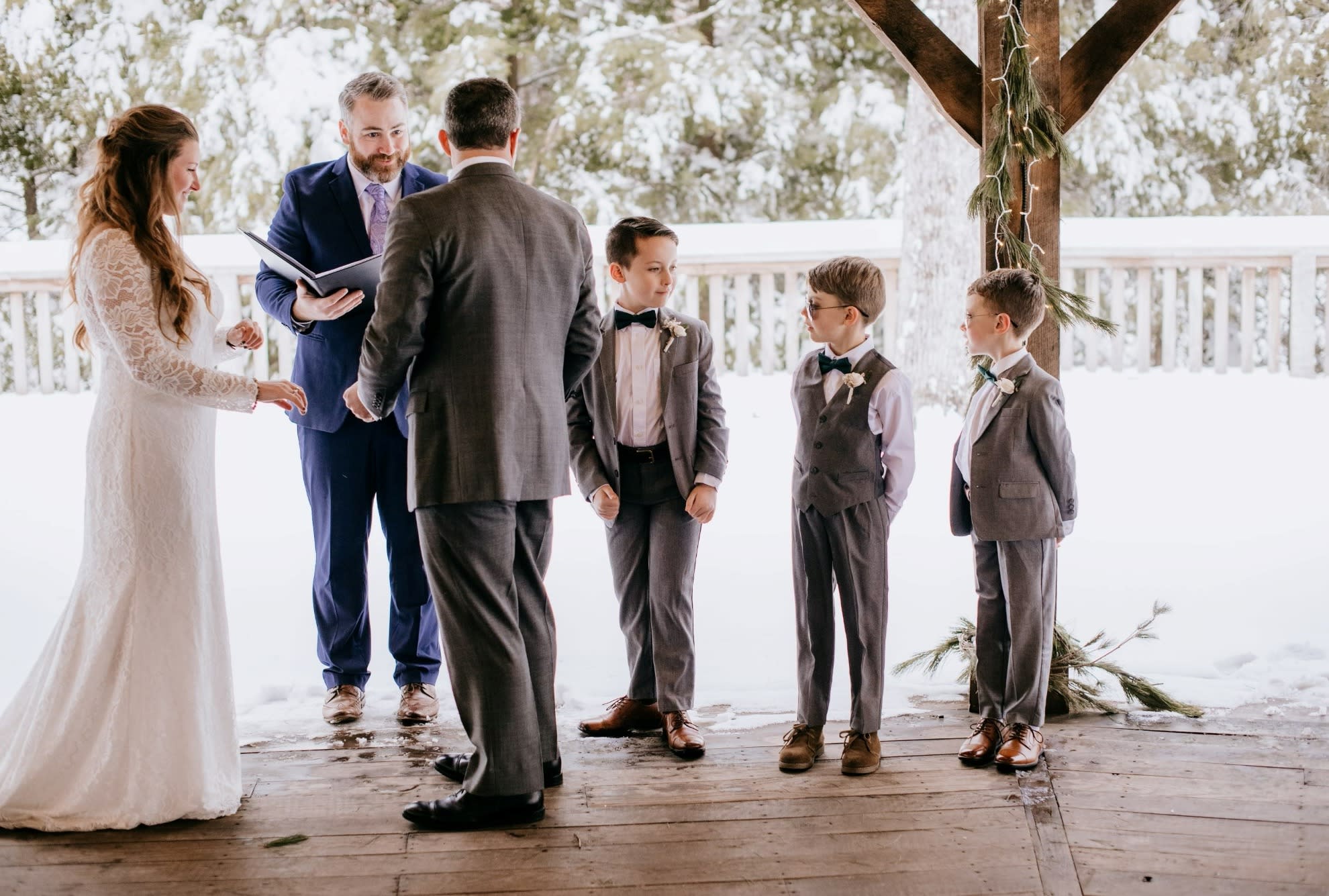 Bride and groom standing under a snow covered pavilion on their intimate wedding day. Three grooms men and a intimate crowd watch as they exchange vows.