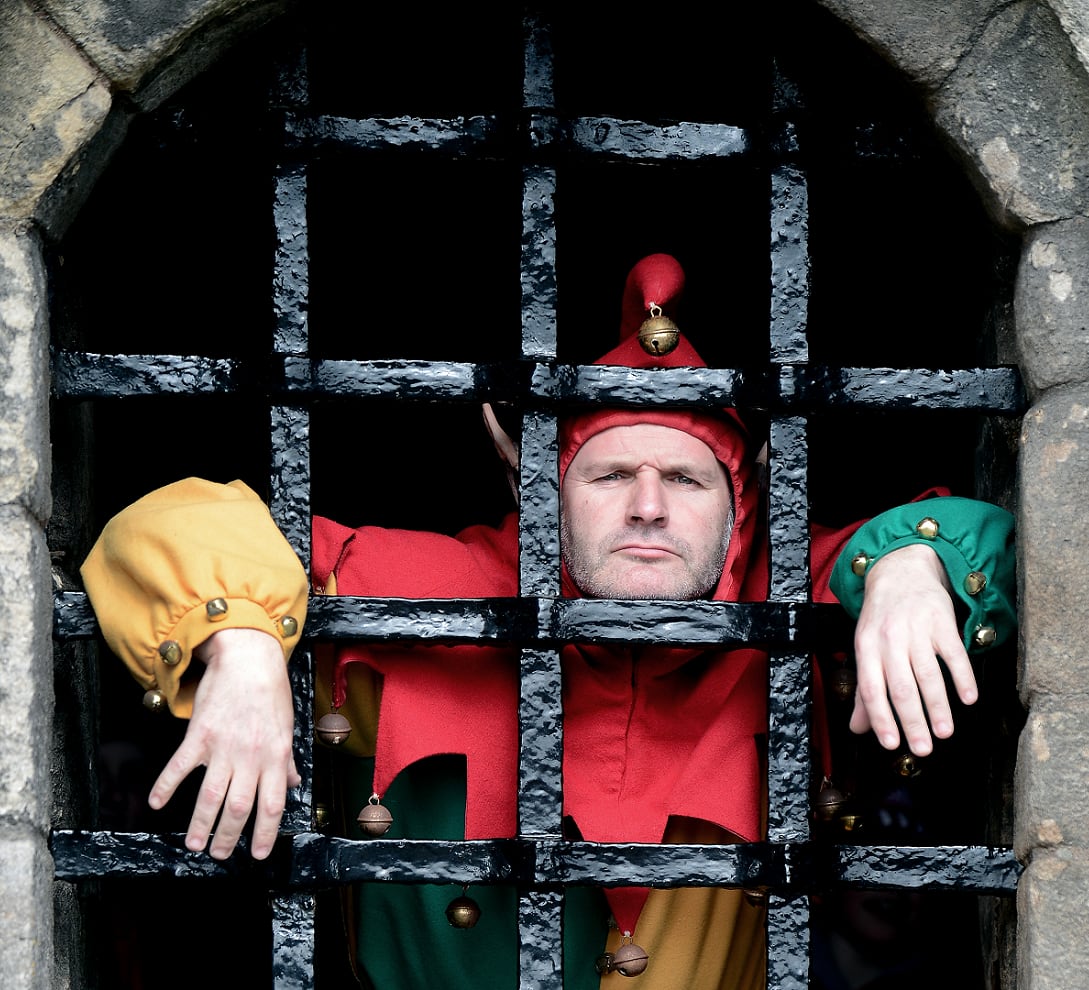 A reenactor dressed as a court jester looking glum as he stands behind bars at a castle