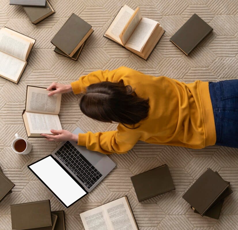 Viewed from above: a woman in a yellow top and jeans lying on her front on the floor, surrounded by books, an open laptop and a cup of coffee, and reading one of the books.