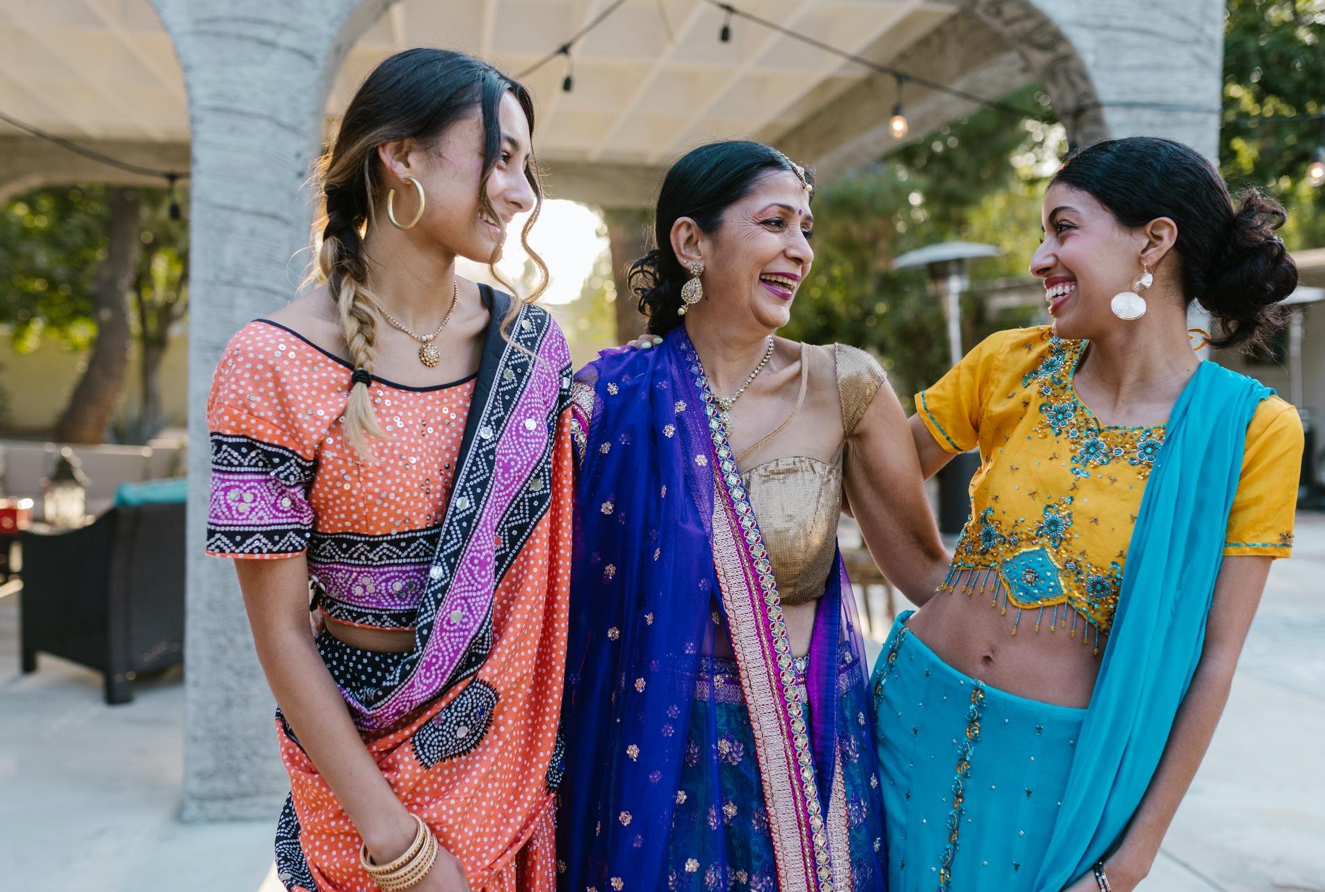 Two young women and an old woman, all in saris, stand together. The youngest-looking one looks at the other two, who are laughing together.