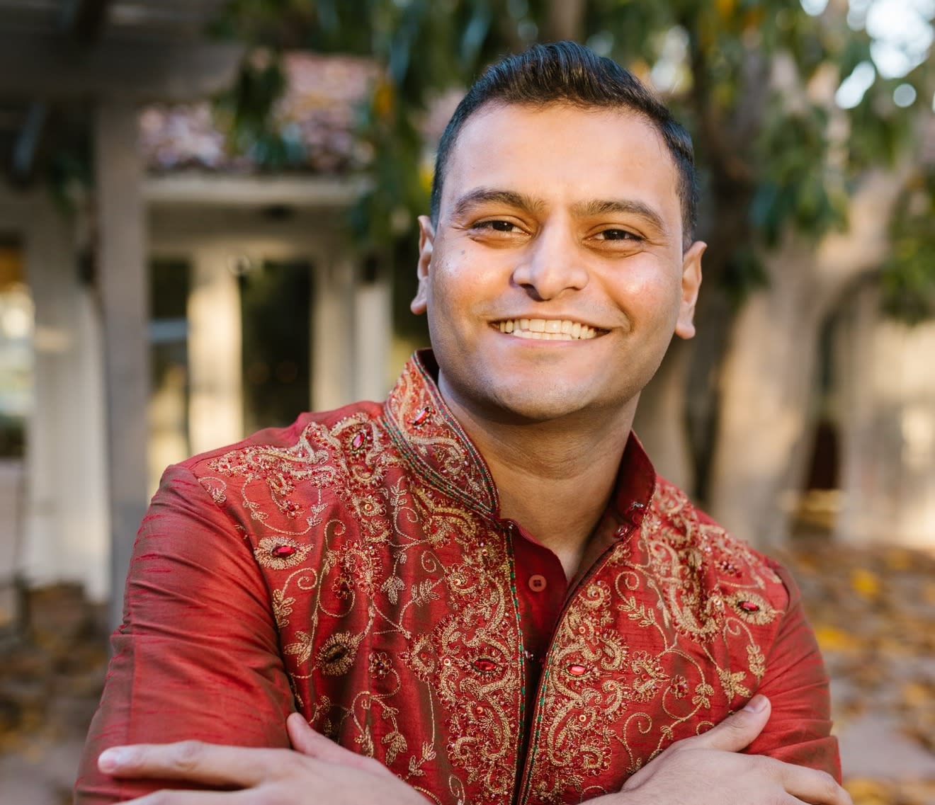 A South Asian man smiles confidently with arms folded over his chest. He's wearing a red kurta heavily embroidered in gold.