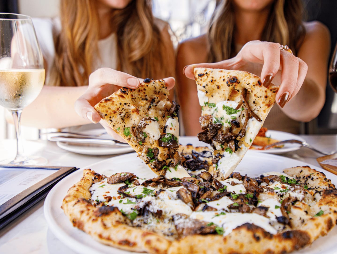 Two women reaching for slices of pizza and drinking wine while out to dinner