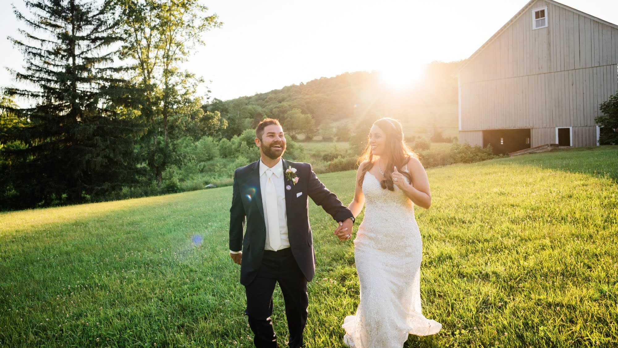 Groom leading bride by the hand away from the barn to a field for golden hour pictures on their wedding day
