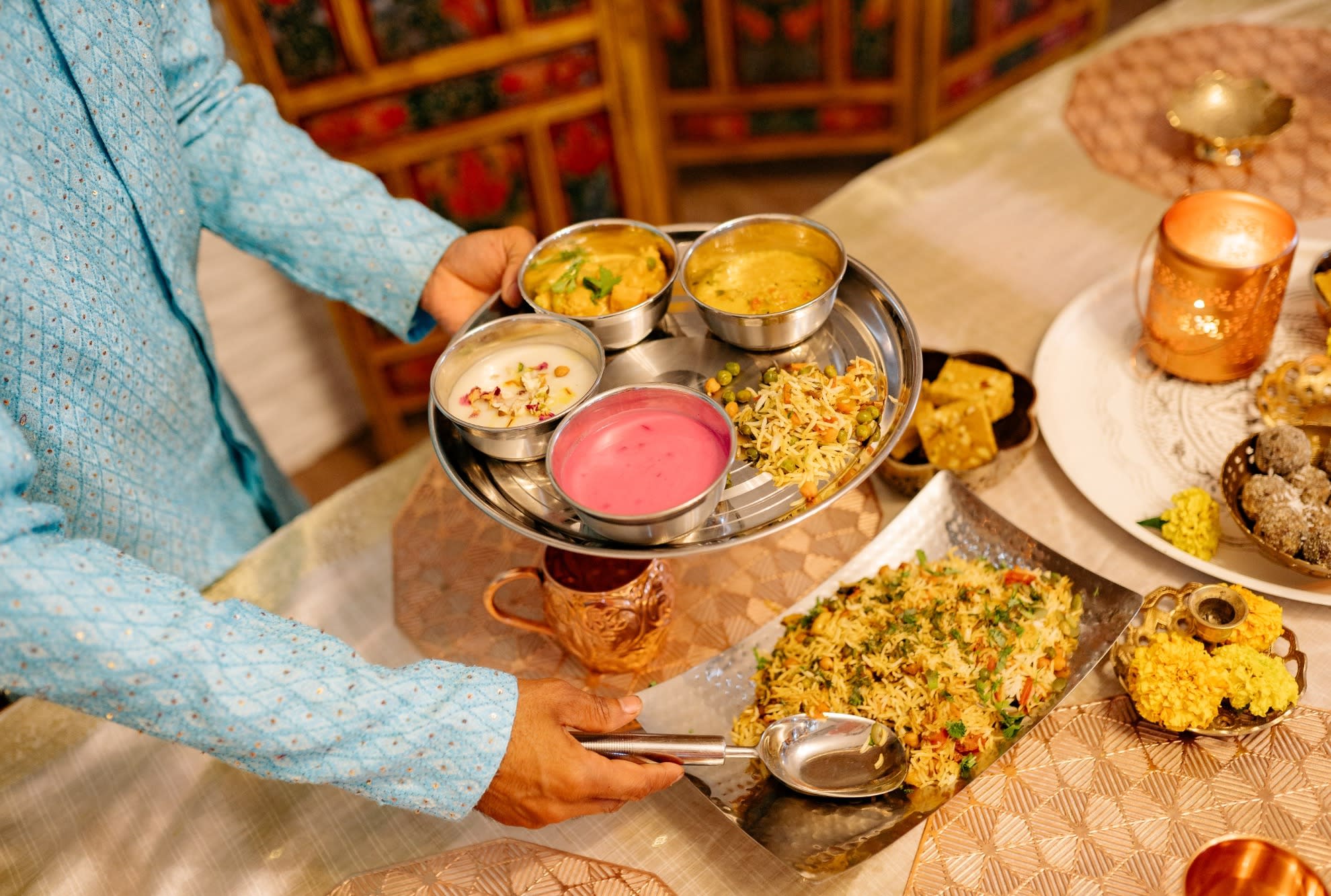 A person in a light blue kurta serves themselves with rice, chutney and yogurt at a richly covered table