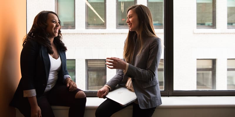 Two business casual women sitting on window ledge giggling