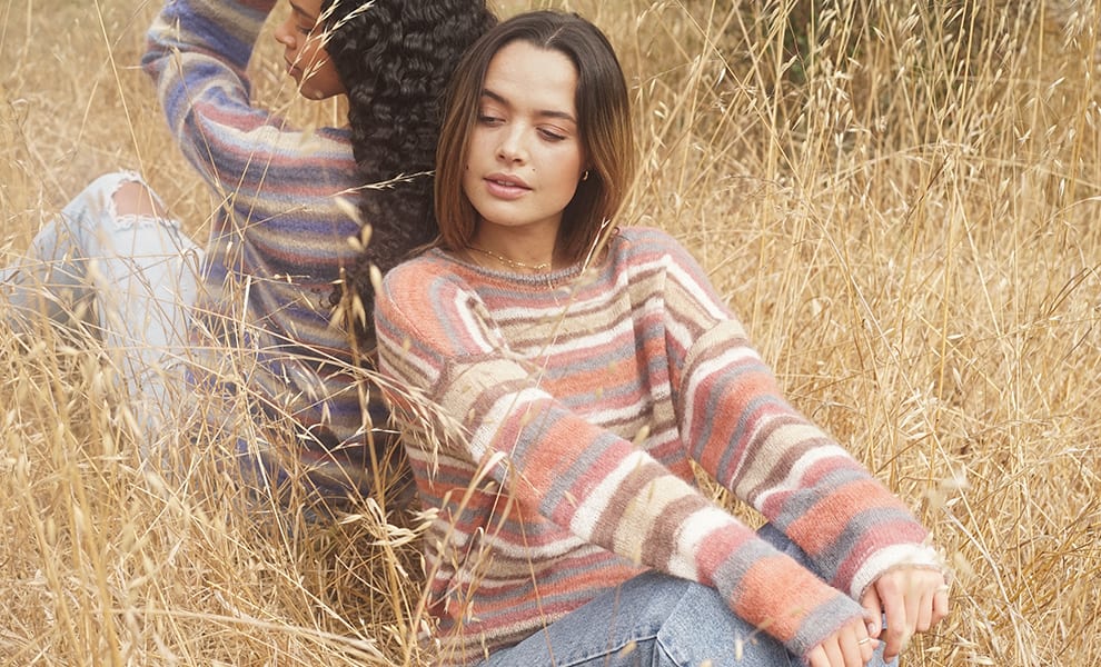 Two women sit back to back in a wheat field, wearing similar striped sweaters.