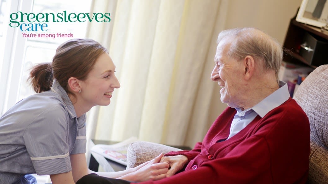 Young female carer looks compassionately at older male resident as she holds his hand