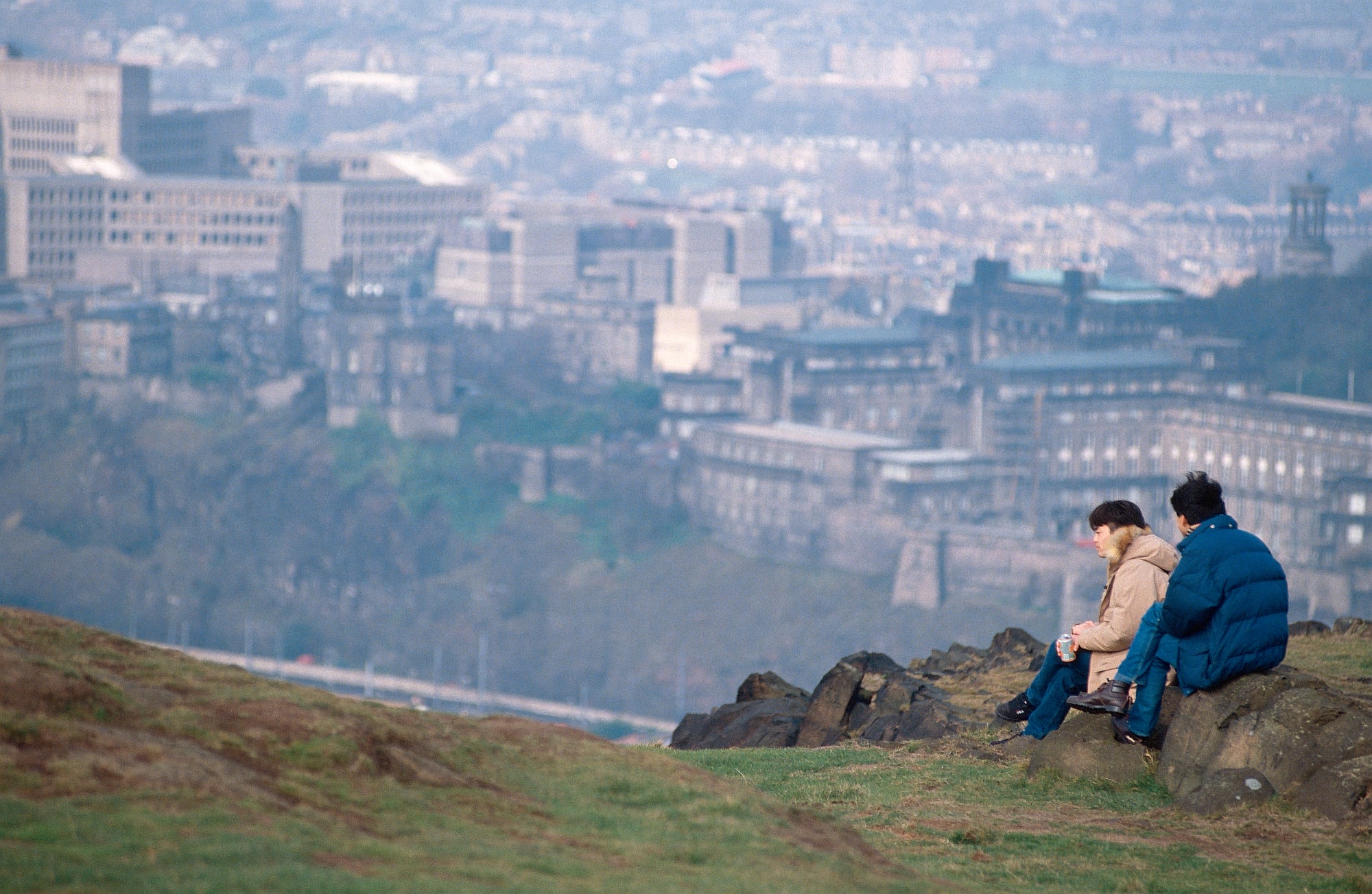 Two people sit on a hill overlooking a city