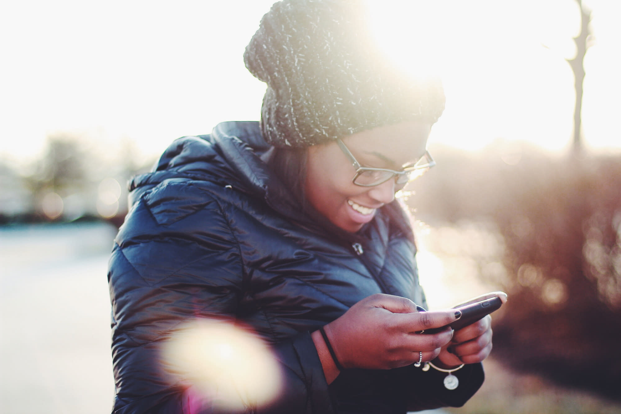Young Black woman with a ribbed bobble hat and black padded jacket on, and glasses, laughing as she texts someone from her smartphone. There are sun glares in the shot.