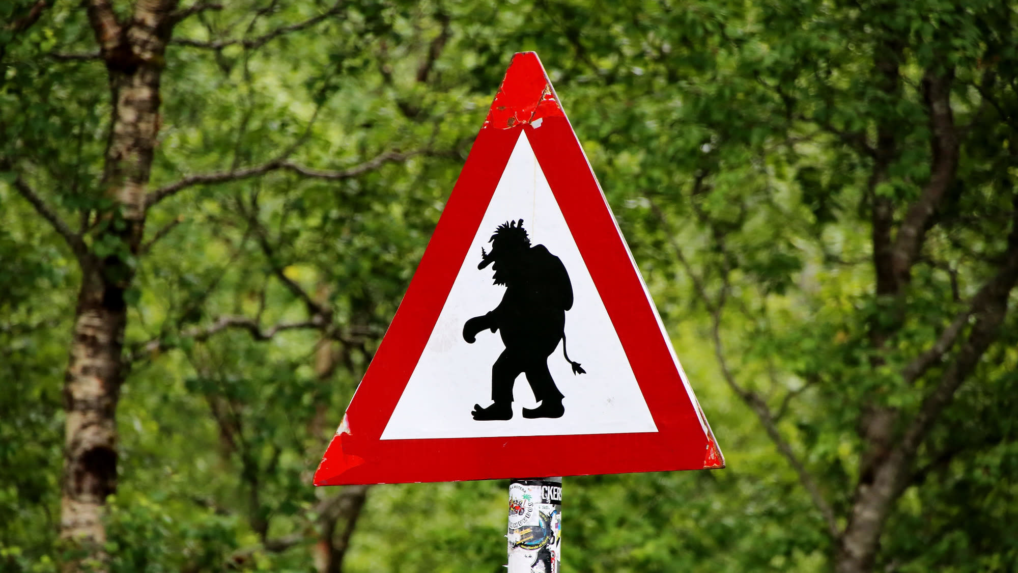 The image of a red triangular Norweigen road sign warning of trolls crossing. There is a background of trees.