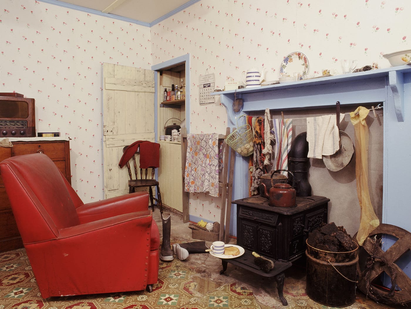 A kitchen with an iron stove, red armchair, blue fireplace and patterned wallpaper